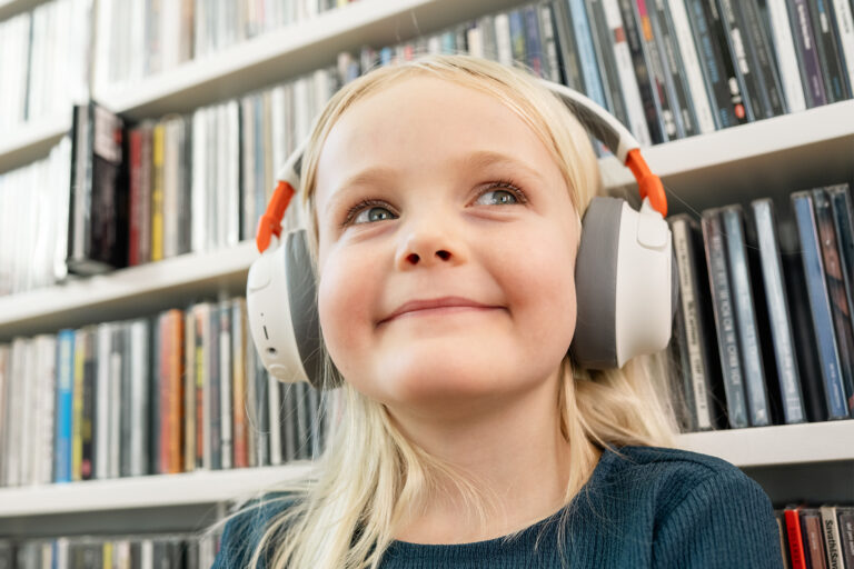 Jente med lyst hår og hodetelefoner. Sitter foran bokhylle, lett smilende.