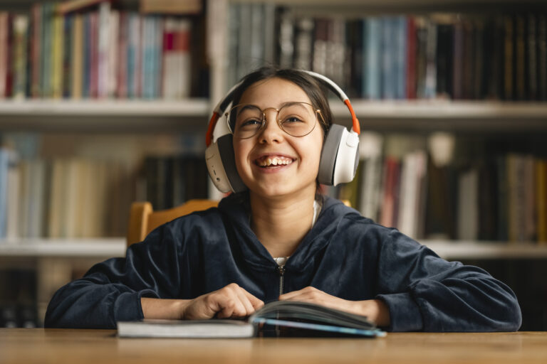 Smilende jente med briller og hodetelefoner sitter og leser i bibliotek med bokhyller.