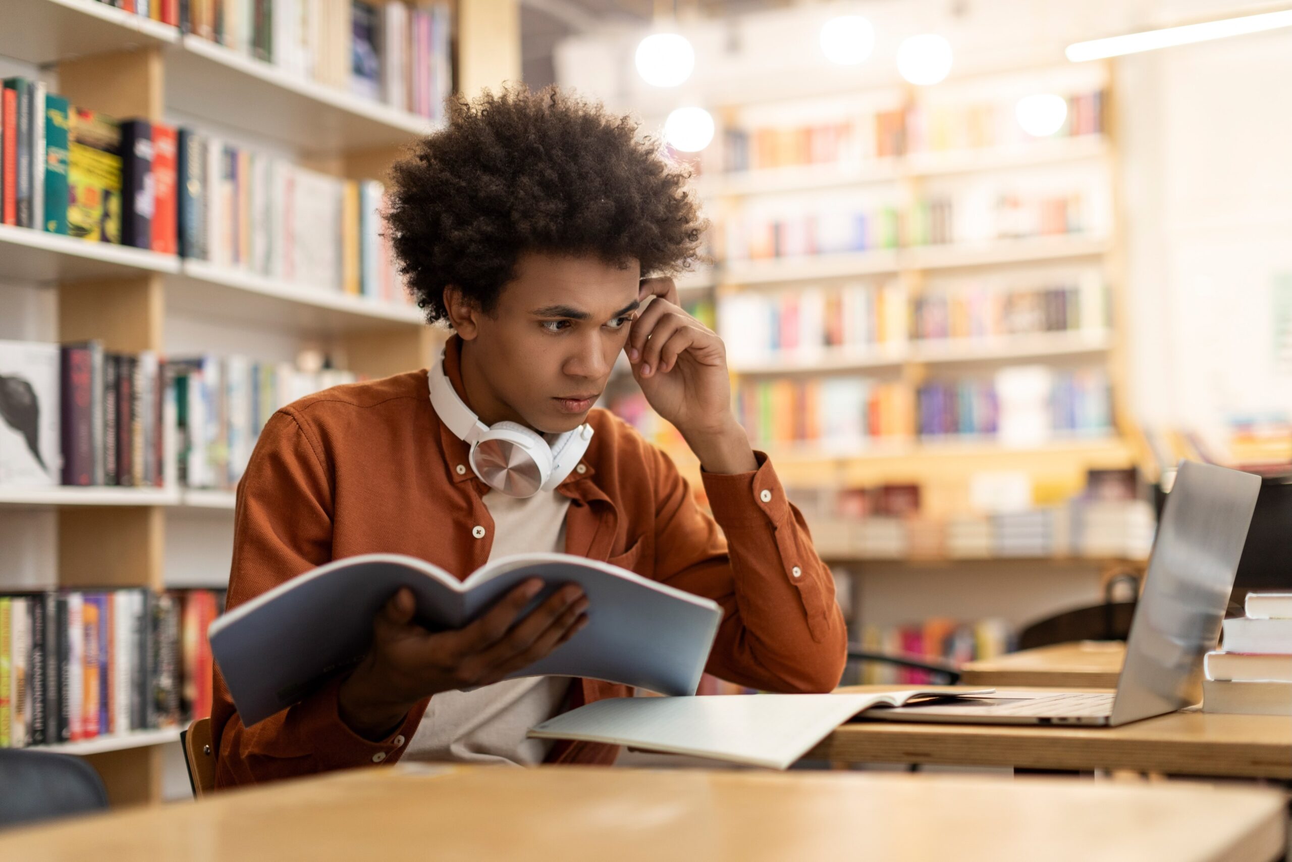 Tankefull mørk, mannlig student i bibliotek med PC, bok og hodetelefoner.