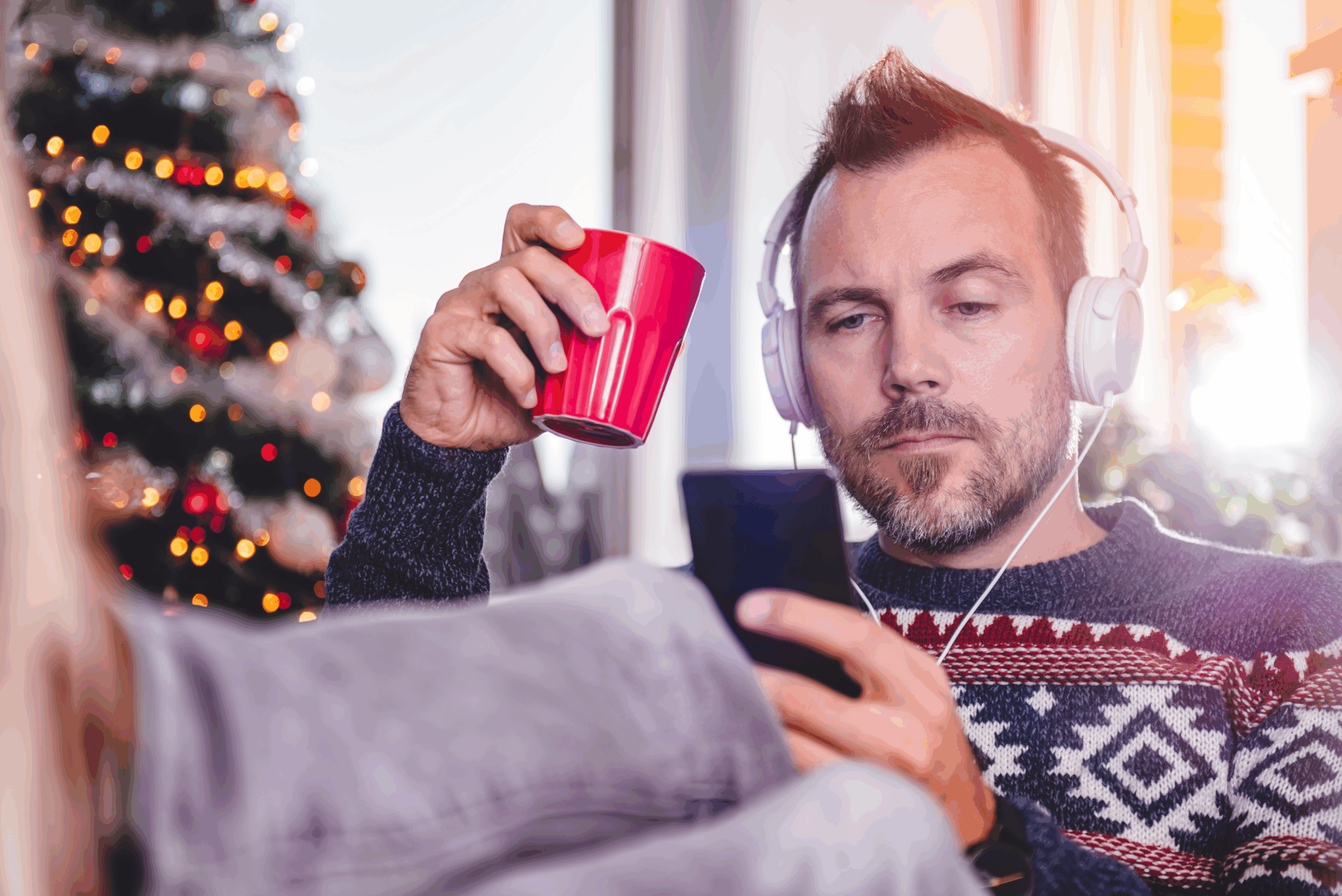 Mann med hodetelefoner, mobil og en rød kopp, med juletre i bakgrunnen.