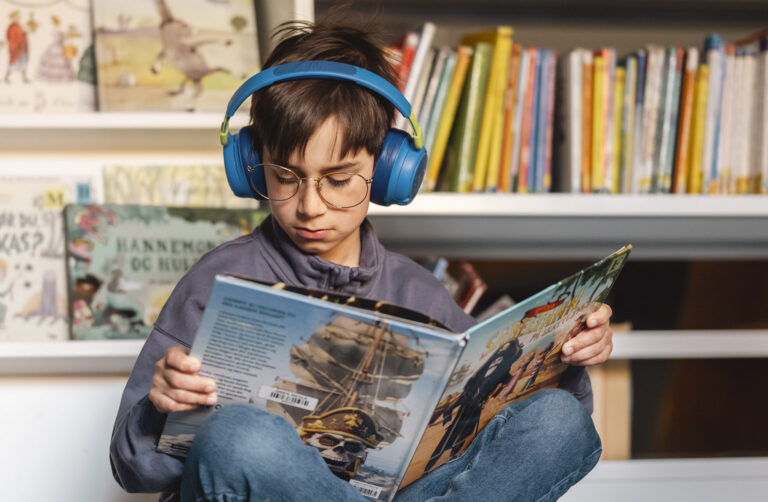 Gutt sitter på gulvet i et bibliotek med hodetelefoner og bok