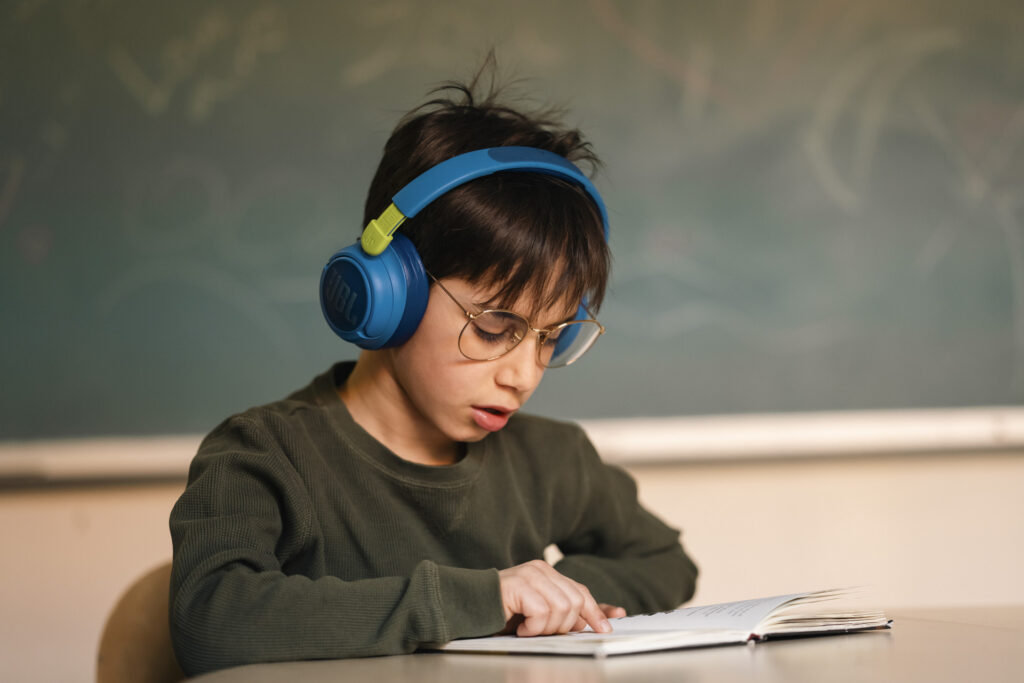 Skolegutt med briller og hodetelefoner sitter ved pulten og leser i en bok. Tavle i bakgrunnen.